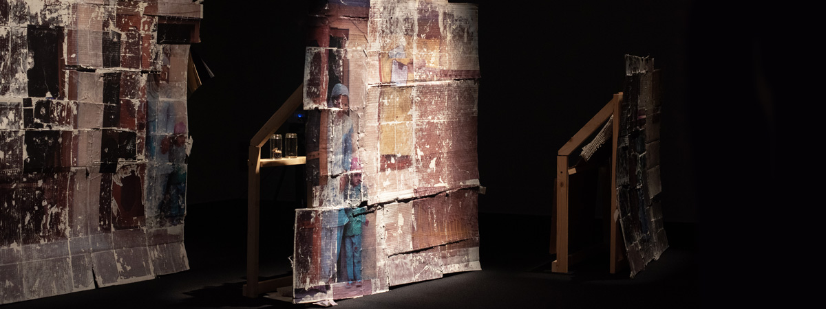 Three structures made of wood and collaged prints in a dimly lit space.