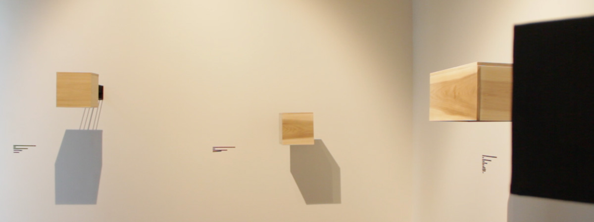 Three wooden boxes mounted on white gallery walls. A black square shape in the right foreground.
