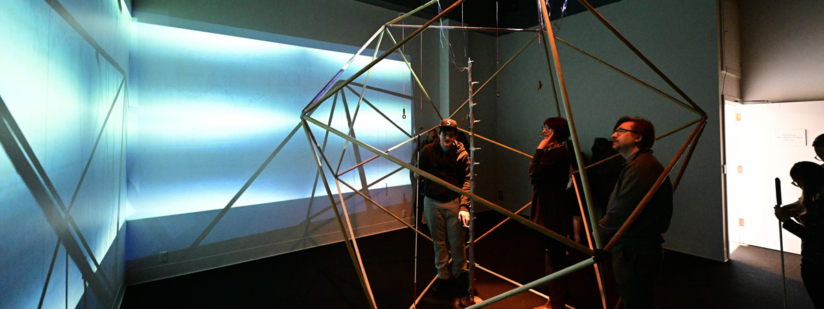 People standing in a large geometric structure made of wooden rods, in a dark gallery space. Abstract images projected on the walls on the left.