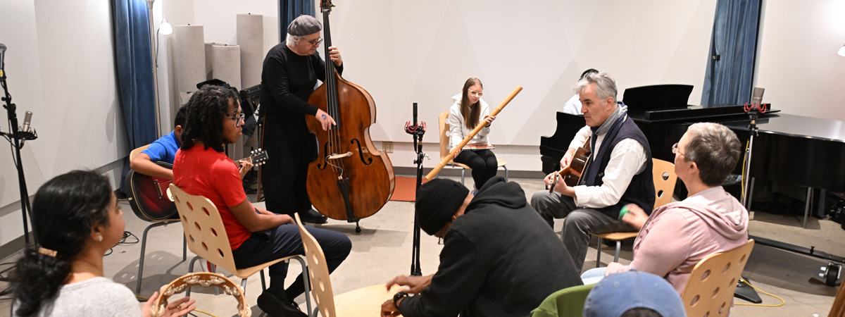 A group of adults and youth play musical instruments in a recording studio, while others listen.