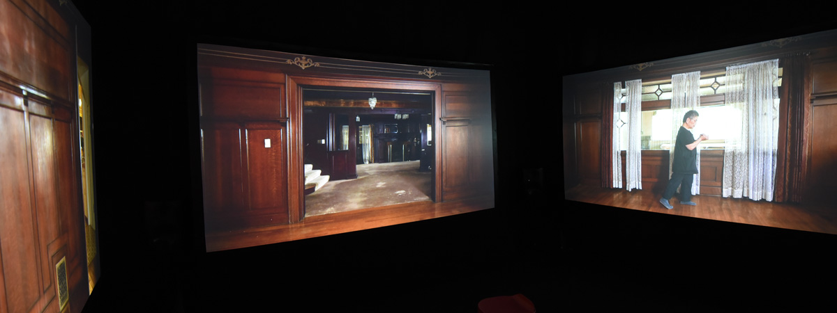Three projection screens on a dark background, showing the interior of a house, with a person dressed in black in front of windows on the rightmost screen.
