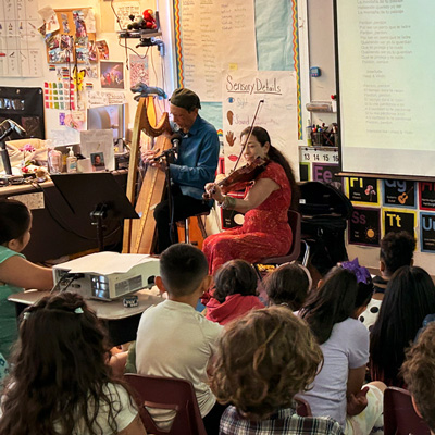 Abel Rocha and Madeleine Sosin playing harp and violin with a classroom of children.