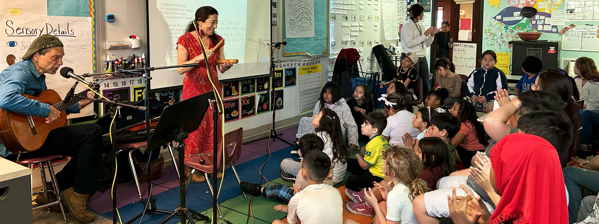 Abel Rocha and Madeleine Sosin playing musical instruments in front of a classroom of students.