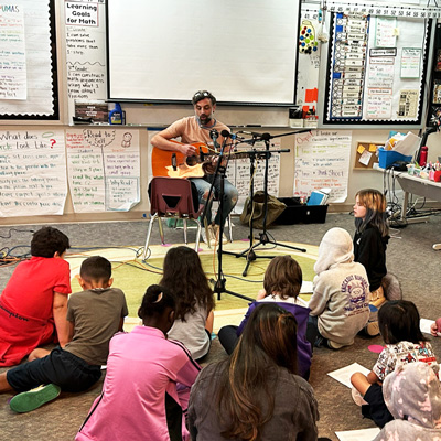 Tito Ramsey seated with a guitar in front of two microphones, with a classroom of children sitting on the floor.