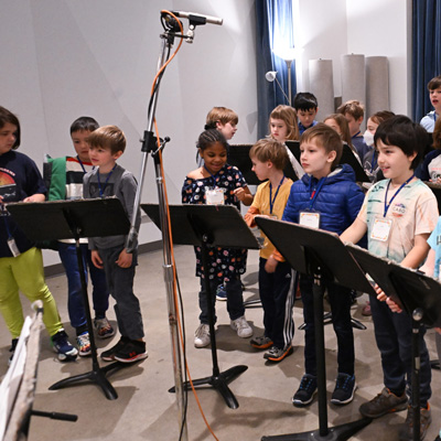 A group of children in a recording studio, with a pair of microphones on a stand above them.