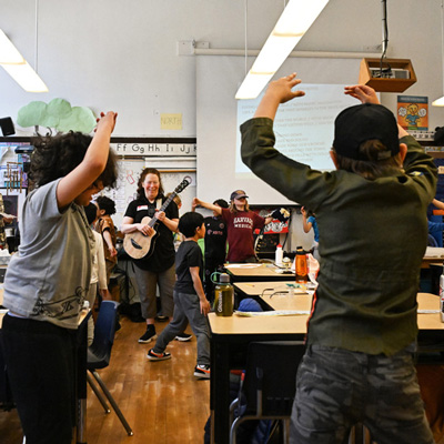 Amy Denio plays guitar while a classroom of children raises their arms.