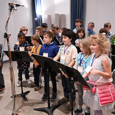 A group of children in a recording studio, with a pair of microphones on a stand above them.