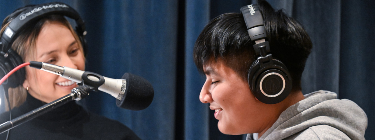 A student at a microphone wearing headphones, smiling, with a vocal coach wearing headphones looking toward him and smiling.