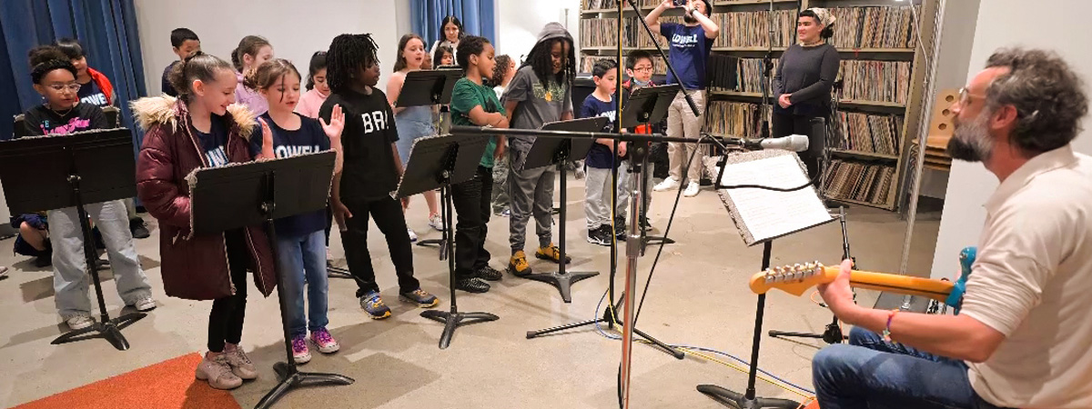 Students singing at music stands while Tito Ramsey, seated, plays guitar and sings.