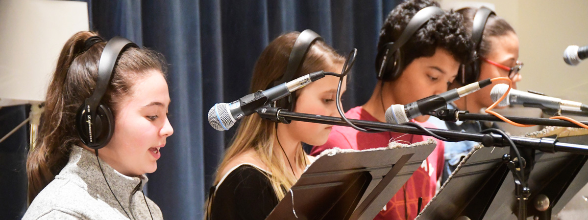 A group of young people wearing headphones, standing at music stands in front of microphones.