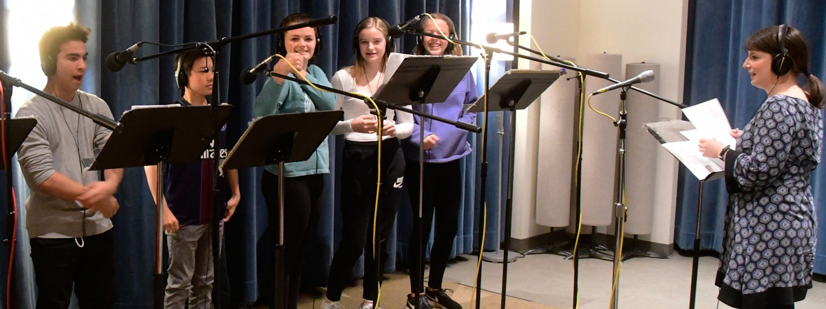 Several young people standing at microphones and music stands, wearing headphones. A woman faces them wearing headphones and holding sheets of paper.