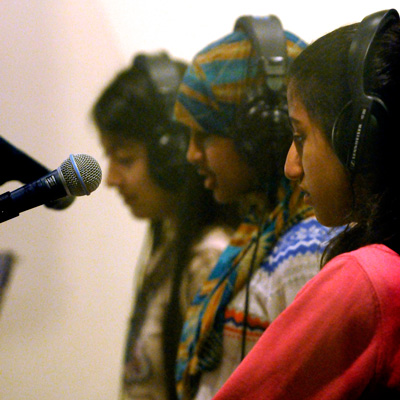 Three people in profile, wearing headphones, facing a microphone.