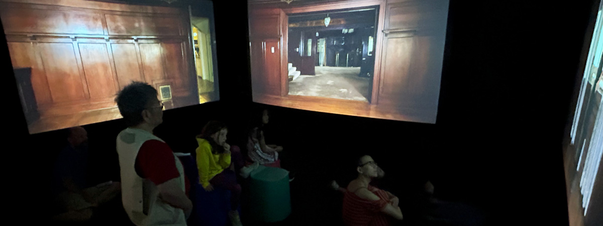 People in a dark room looking at three projection screens, showing the interior of a house.