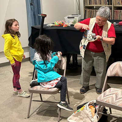 Susie Kozawa operates a hand-cranked instrument while two youth look on, one of them holding a tin can. Other objects in the background.