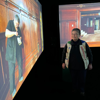 Susie Kozawa in a dark space with two projection screens showing the interior of a wooden house. Susie appears on the lefthand screen, in the foreground.