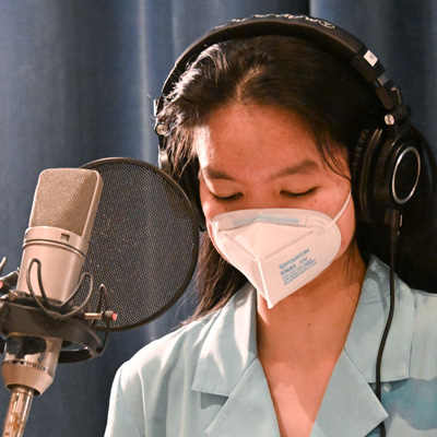 Allison Masangkay, wearing headphones and a KN95 mask, standing at a microphone.