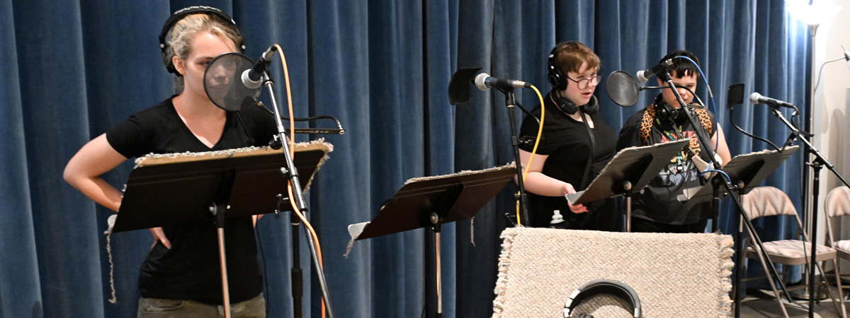 Three people standing at music stands in front of microphones, wearing headphones.