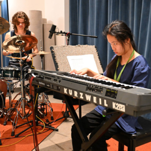 Two people in a recording studio, playing drum kit and synthesizer