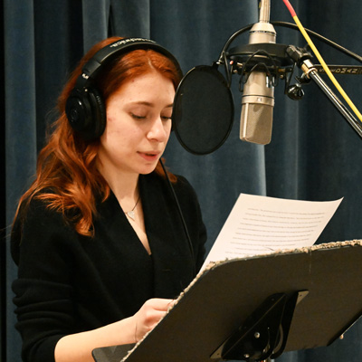 McKenna Princing wearing headphones, standing at a microphone, reading from a piece of paper.