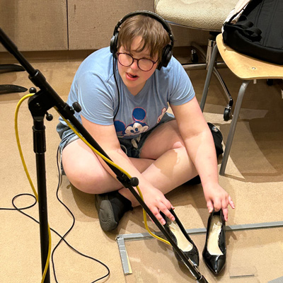 A person seated on the floor holding a pair of shoes on a sheet of clear plastic, with a microphone pointing toward the shoes.