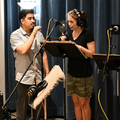 Two people in a recording studio. One stands at a microphone with headphones, while the other holds his hand out from his face with thumb and pinky extended.