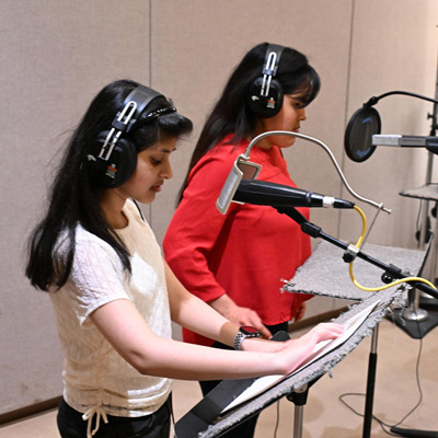 Two people wearing headphones standing at microphones. One holds her hands on a sheet of white paper on a music stand.