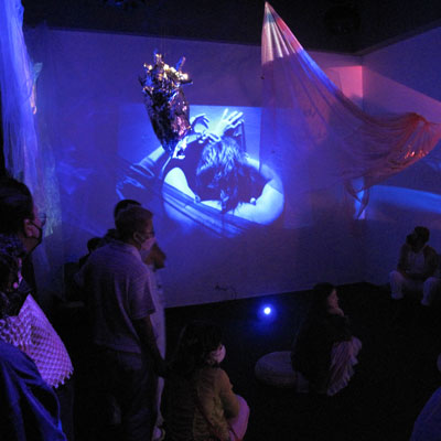 People in a dark gallery space with projections on the walls and hanging fabric and sculptures.