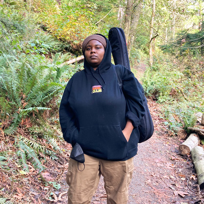 Rasheena Fountain, standing on a wooded path, holding a guitar in a gig bag.