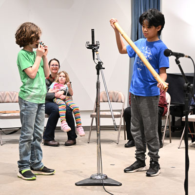 Kids standing around a microphone, playing instruments.